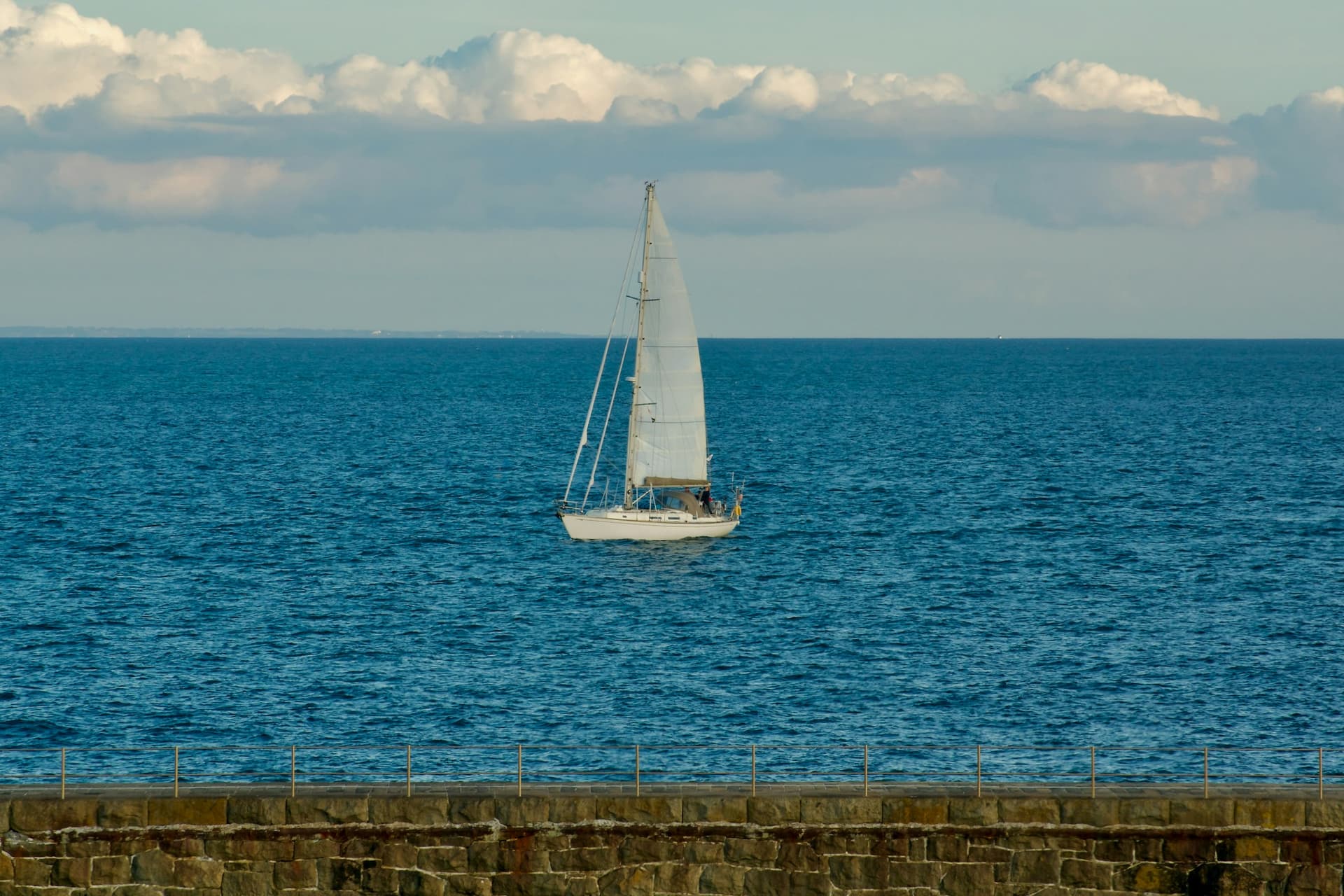 A white cruising sloop under sail on deep blue water, beneath a bank of white cloud.
