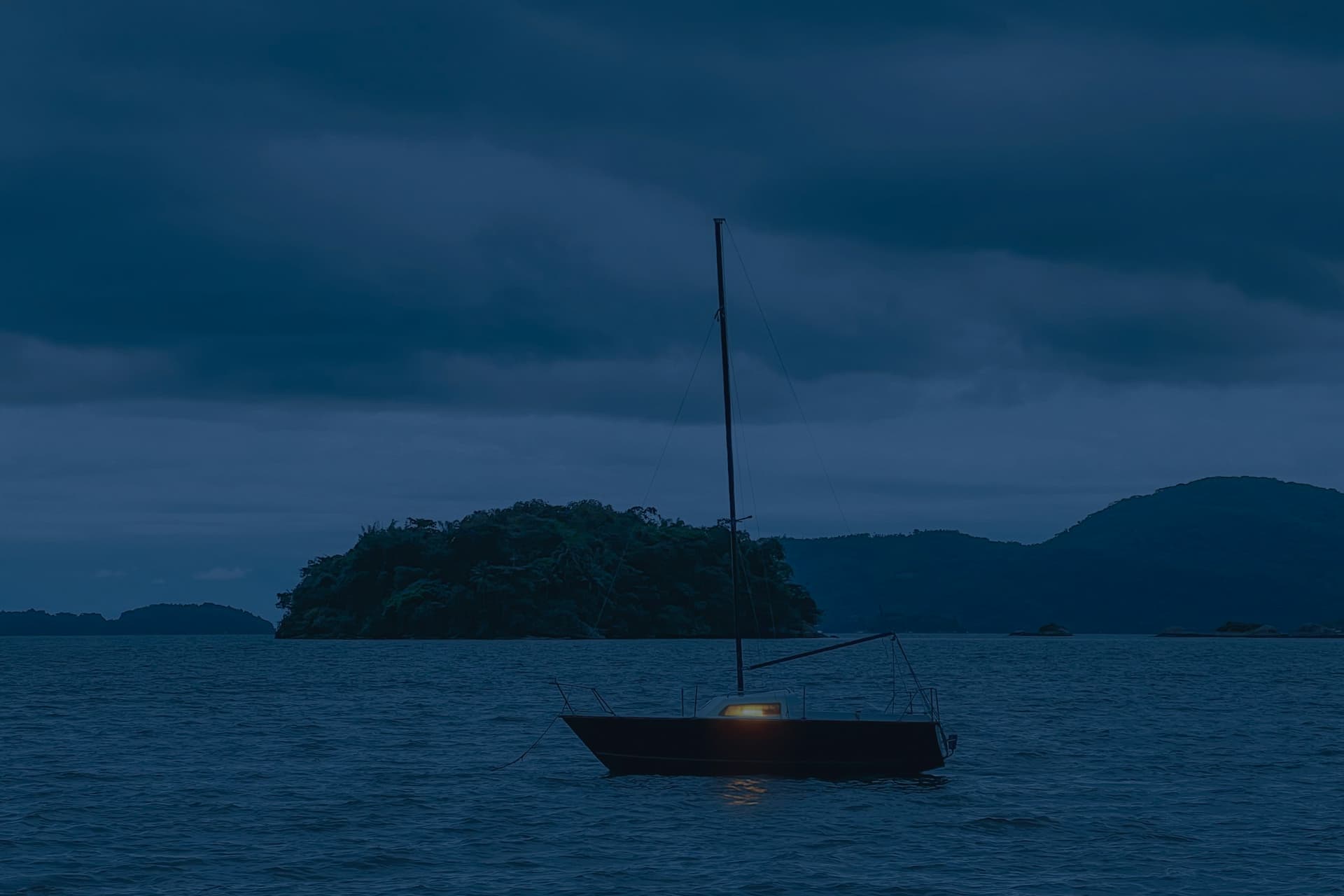 A small sailboat at anchor at dusk under heavy blue cloud, one cabin light lit, a wooded island behind it.