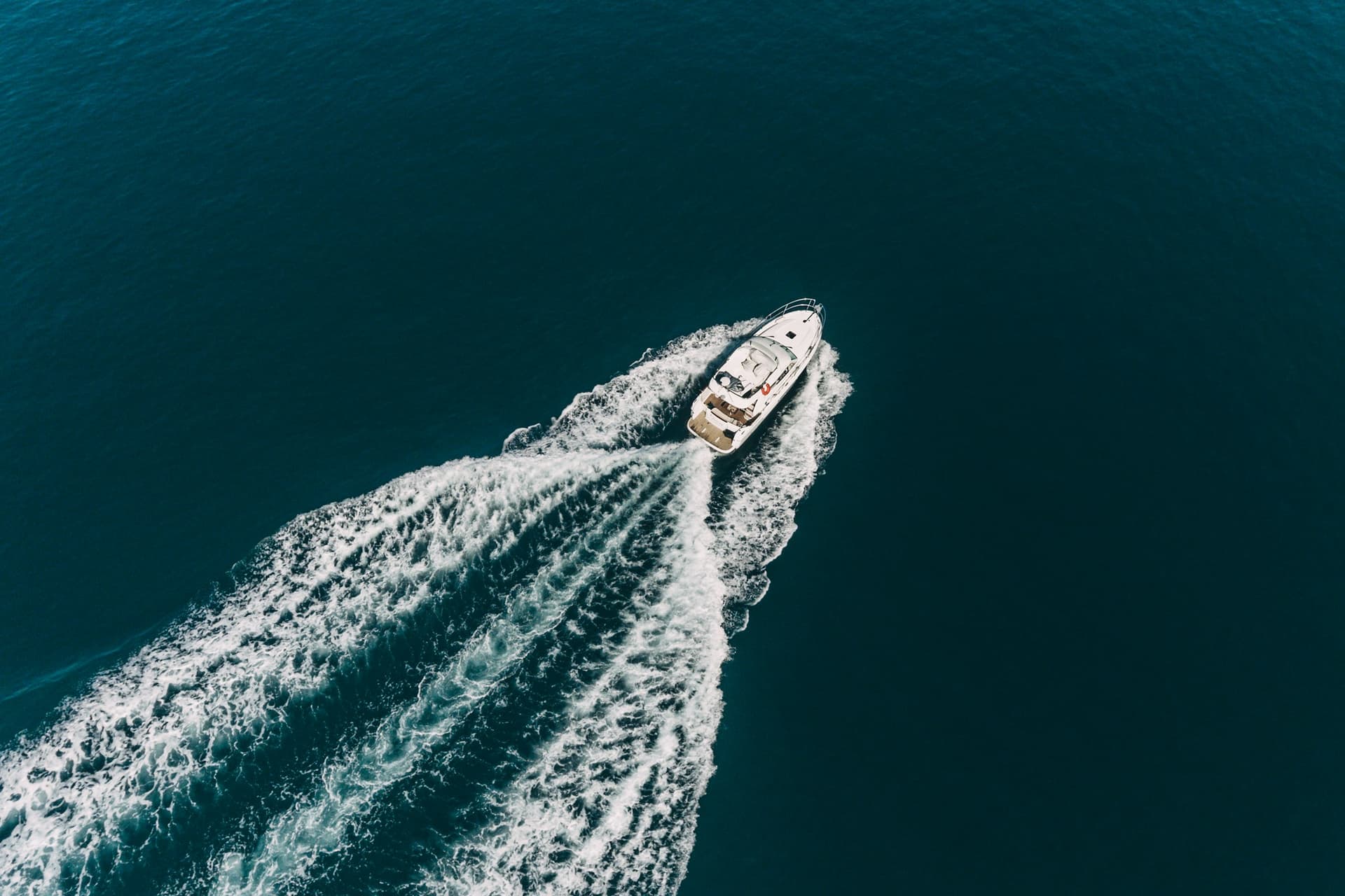 A white motor cruiser seen from above, running across deep blue water with a wide white wake behind it.