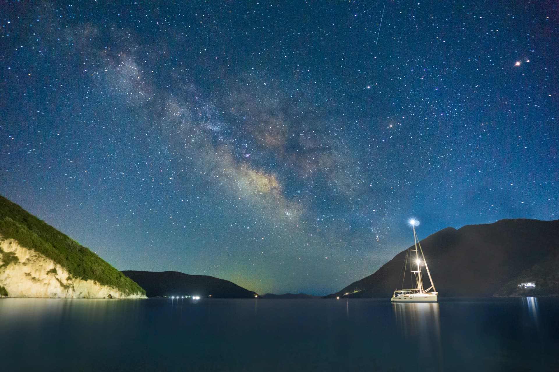 A sailboat at anchor in a calm bay under the Milky Way, a white light shining at the top of its mast.