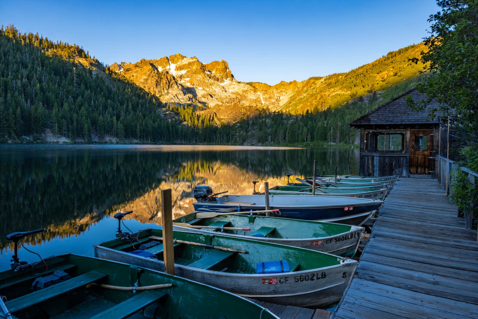 Small aluminum skiffs with trolling motors and an outboard, tied up at a wooden dock and boathouse on a mountain lake at sunrise.