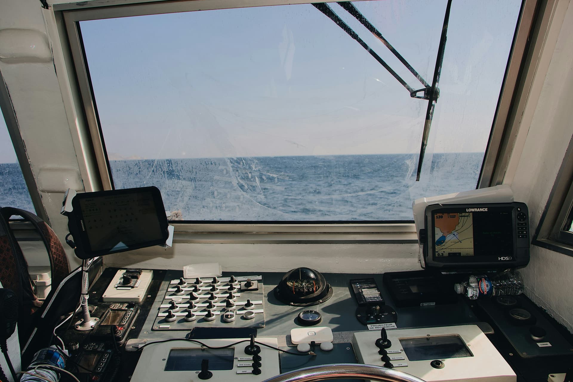 A boat's wheelhouse looking out over the bow to open sea, with a chartplotter showing a chart, a compass and the helm controls.