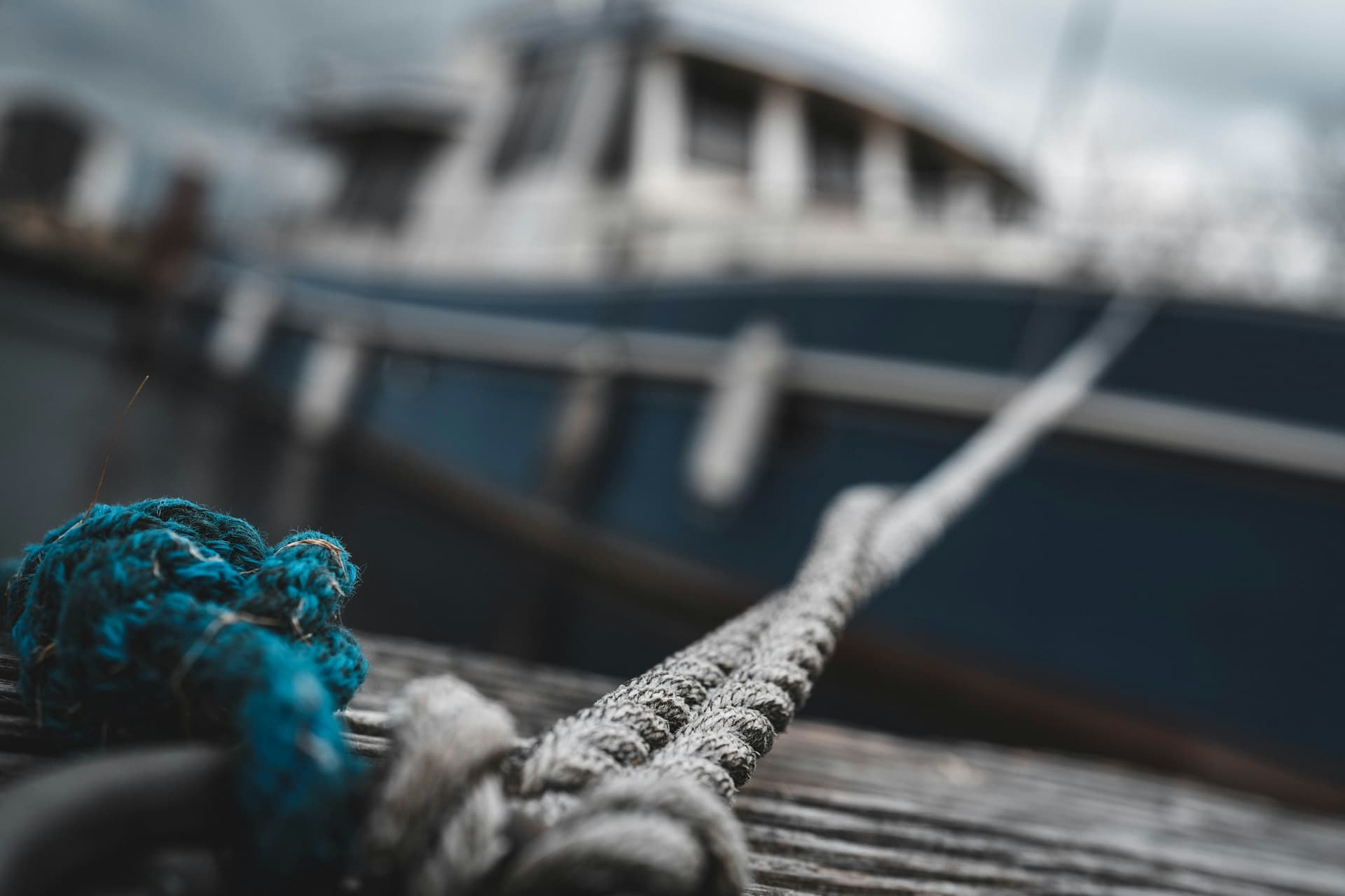 A taut grey line lying across a wooden dock toward a blue-hulled boat with white fenders hanging along its side, with a coil of blue rope in the foreground.