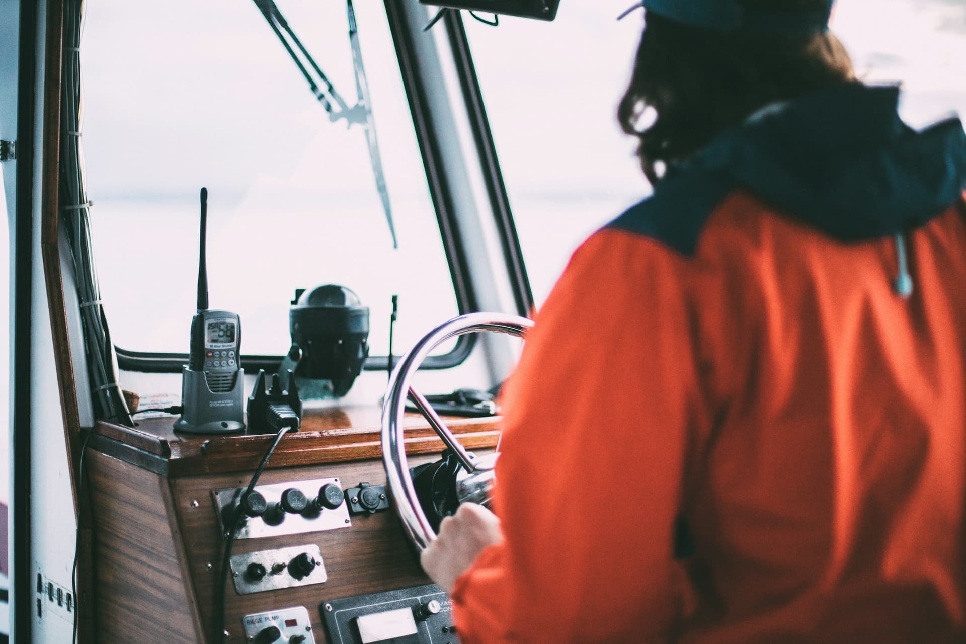 A handheld marine VHF radio in its charging cradle on a boat's dashboard, beside the compass, with the skipper at the wheel.