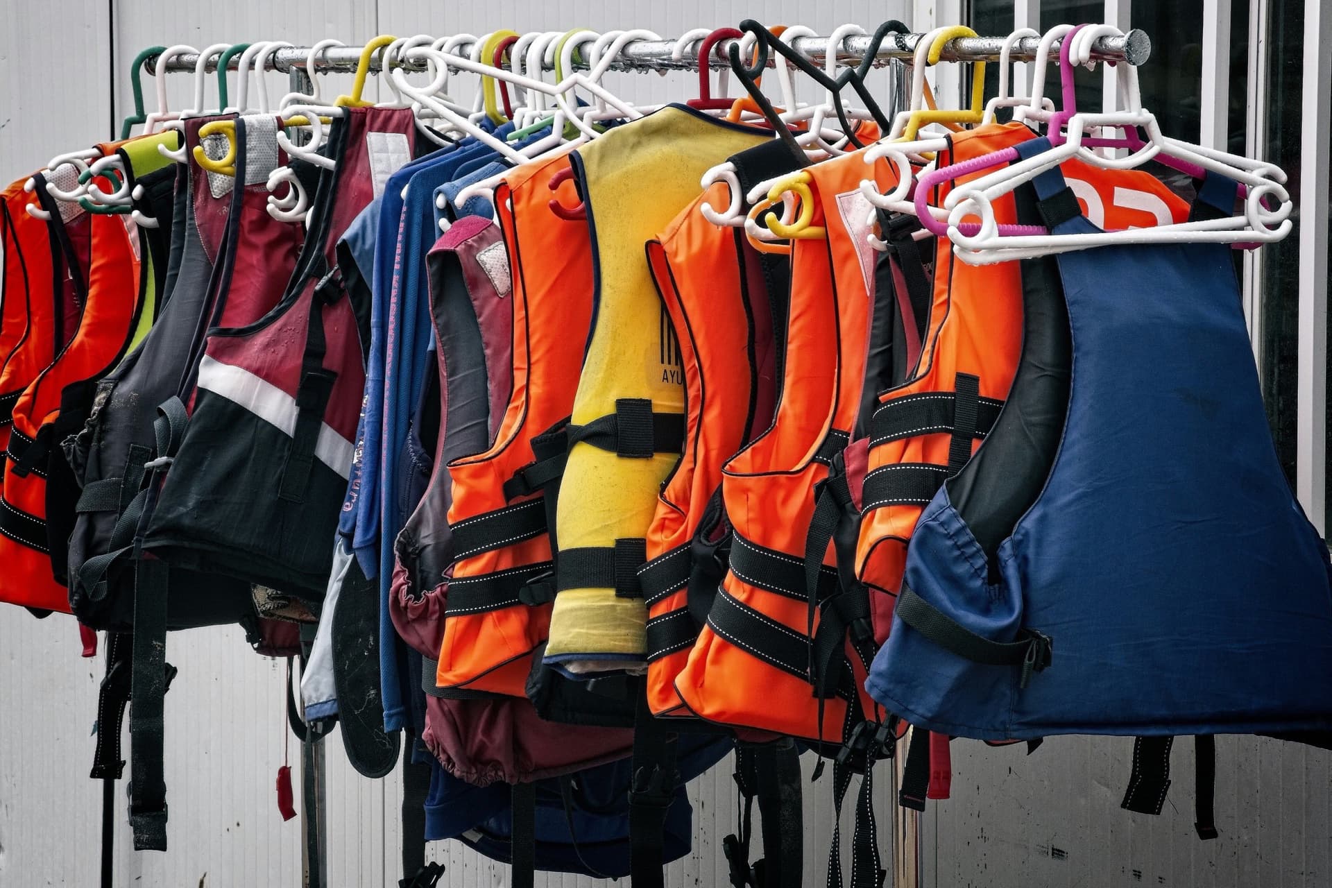 Foam life vests in orange, yellow, red, navy and blue hanging from a rail on plastic hangers.