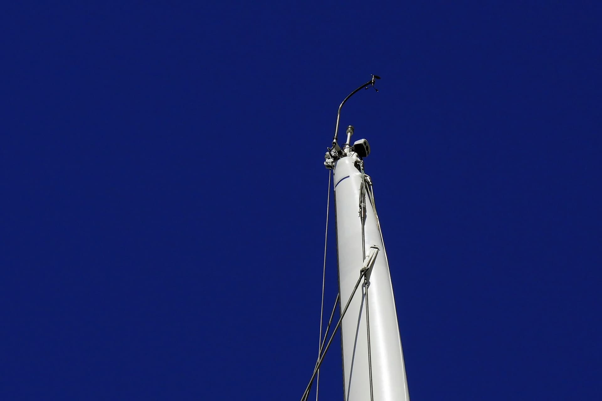 The top of a white sailboat mast against a deep blue sky, with a wind sensor on a curved arm at the masthead.